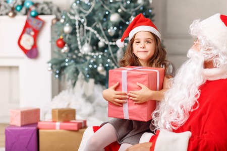 Merry Christmas. Nice little girl holding present and sitting with Santa with Christmas tree standing in the backgroundの写真素材