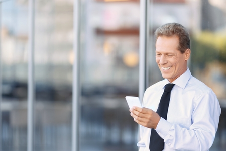 modern businessman. Handsome positive businessman holding mobile phone and standing near office building while going to make a callの写真素材