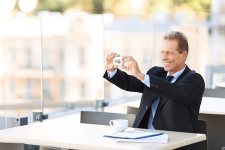 Just smile. Pleasant upbeat handsome businessman holding camera and sitting at the table while making photosの写真素材