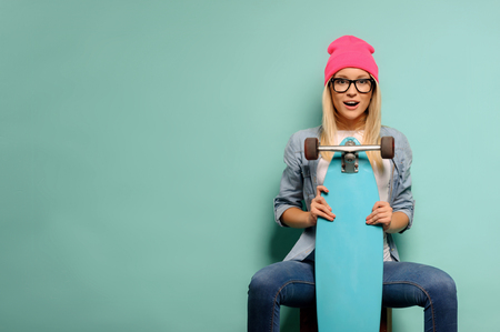 On the edge of emotions. Pleasant upbeat young girl holding her skateboard and sitting while being isolated on blue backgroundの写真素材