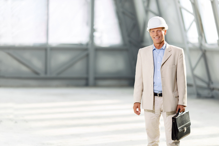Real professional. Pleasant smiling cheerful delighted architect holding case and wearing helmet while workingの写真素材