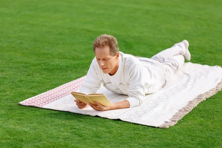 Best one. Pleasant positive adult man lying on the blanket and holding book while reveling in itの写真素材