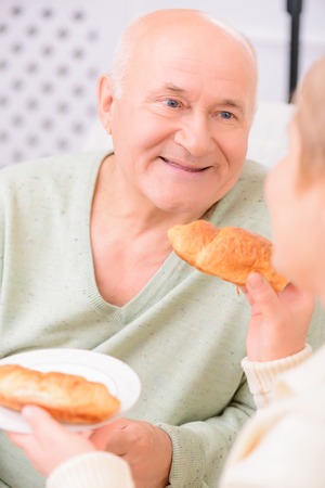 So tasty. Close up of pleasant adult content man sitting in the arm chair and eating croissants while looking at his wifeの写真素材