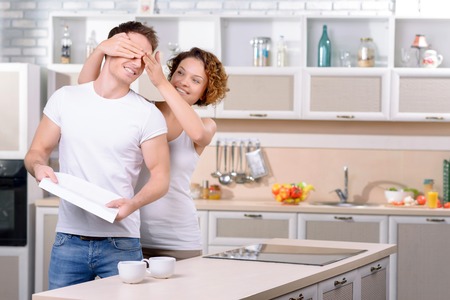 Guess who. Agreeable young vivacious girl closing eyes of her boyfriend and evincing joy while standing in the kitchenの写真素材