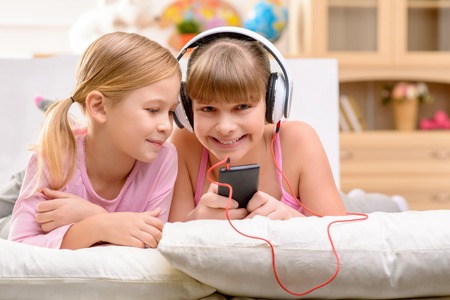 Pleasant smiling little girl holding mobile phone and listening to music in headphones while lying on the sofa with her sisterの写真素材