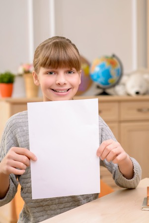 Nice smiling joyful little girl holding sheet of paper and expressing positivity while sitting near the tableの写真素材