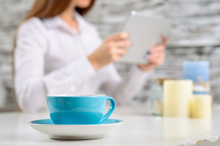 Close up of cup with coffee standing on the table with nice attractive girl sitting in the background and holding tabletの写真素材