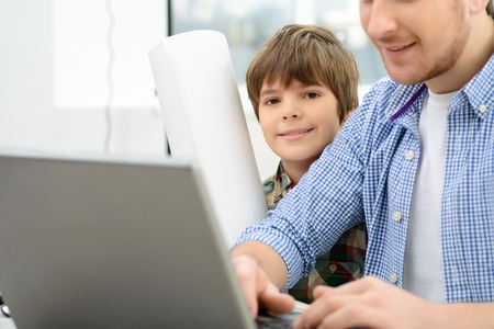 In a good mood. Cheerful delighted little son sitting with father at the table and surfing the...