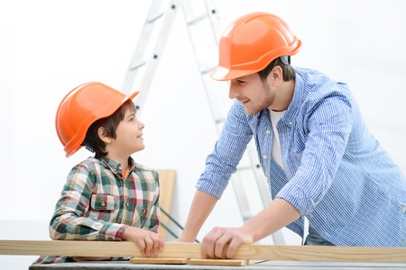 Make everything yourself. Pleasant cheerful father and his son holding stick of wood and smiling while doing renovation togetherの写真素材