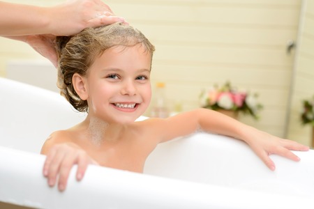 Full of delight. Pleasant little girl sitting in the bath tube and smiling while her mother washing her hairの写真素材