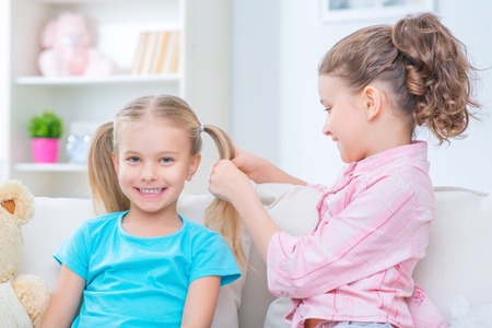 Happy together. Upbeat cheerful little girl making hairstyle to her sister while playing on the sofa and feeling contentの写真素材