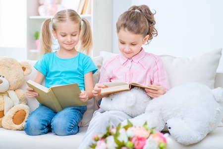 Broaden your mind. Smart little sisters sitting on the sofa and reading while feeling cheerfulの写真素材