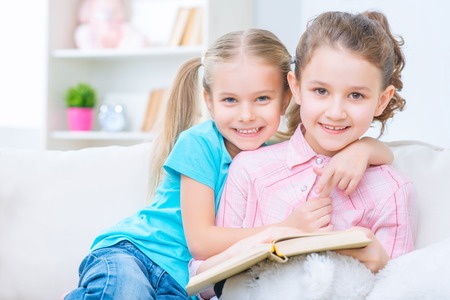 Forever together. Pretty upbeat little sisters embracing and sitting on the sofa while reading  bookの写真素材