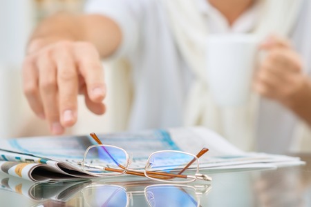 Bad eyesight. Selective focus of glasses lying on the table with senior man holding hand to get them while drinking tea on the sofaの写真素材