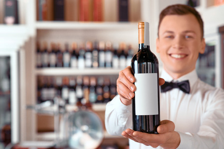 You will like it. Close up of wine bottle in hands of professional positive handsome sommelier holding it while offering it for youの写真素材