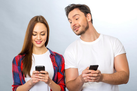 Have a glance.  Young positive couple holding mobile phones while standing isolated on grey...