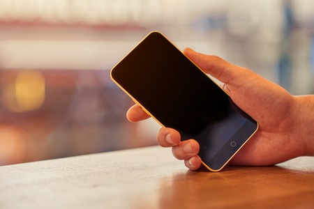 Contemporary life. Close up of smart phone in hands of a pleasant man holding it while sitting at the tableの写真素材