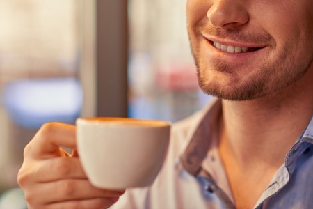 Feel the taste. Cheerful handsome man smiling and holding cup of coffee and drinking it while...