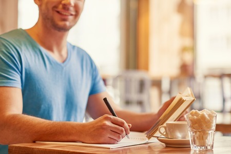 Full of joy. Delighted handsome man sitting at the table and making notes while drinking coffeeの写真素材