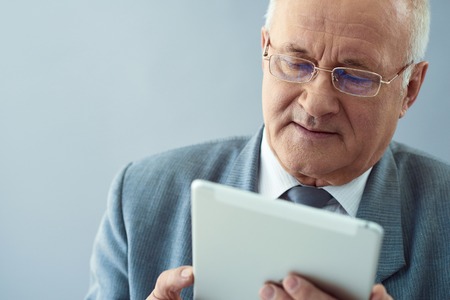 Business plan. Close up shot of wise old man holding tablet and looking thoughtful isolated in grey background.の写真素材