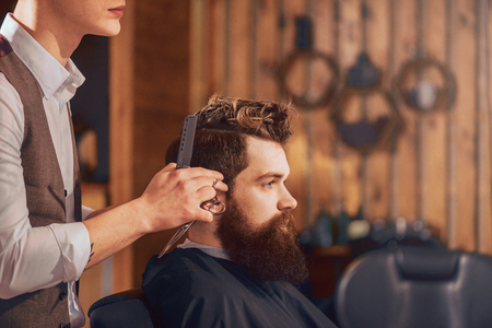 Devoted worker. Bearded handsome man sitting in the barbershop while professional barber making him a haircutの写真素材