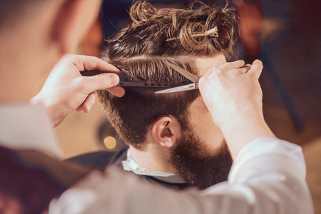 Be accurate. Pleasant bearded man sitting in the barbershop while professional barber making him a haircut with scissors.の写真素材