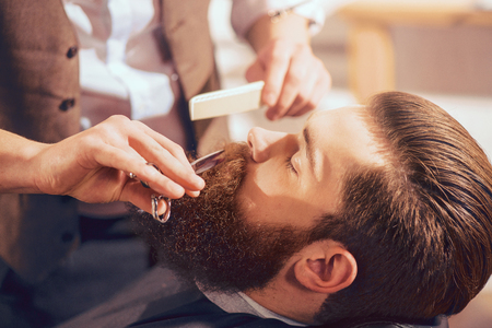 Crafty hands. Handsome man sitting in the barbershop  while professional barber holding scissors and cutting his beardの写真素材