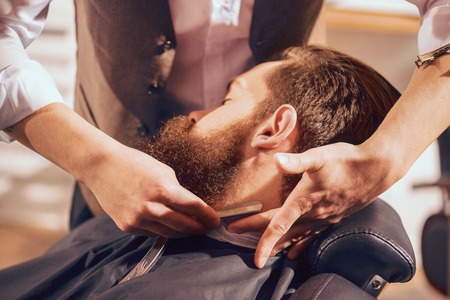 Creative craft. Pleasant professional barber holding special device while shaving beard of his clientの写真素材