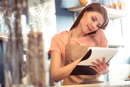 Important call. Pleasant beautiful waitress talking on mobile phone and making notes while being busy at workの写真素材