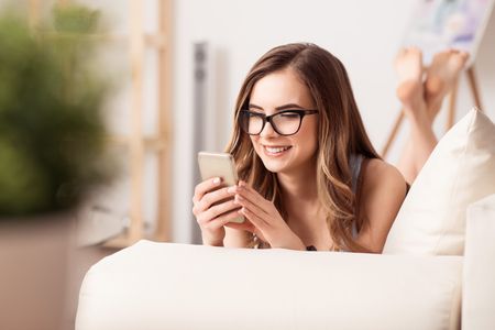 Involved in chat. Cheerful positive beautiful girl holding mobile phone and chatting while lying on the sofaの写真素材