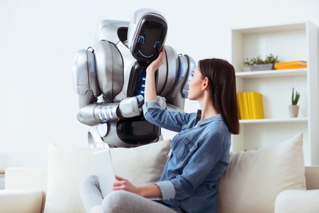 Just smile.  Delighted attractive positive girl sitting on the couch and touching robot while looking at itの写真素材