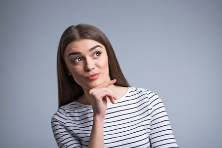 Thoughtful mind. Positive beautiful young girl holding her hand under the chin and looking aside while thinkingの写真素材