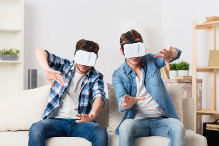 Open your world. Cheerful delighted handsome guys sitting on the couch and having fun while using virtual reality deviceの写真素材
