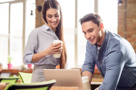A little relaxation. Cheerful positive delighted smiling colleagues drinking coffee and using laptop while resting in the officeの写真素材
