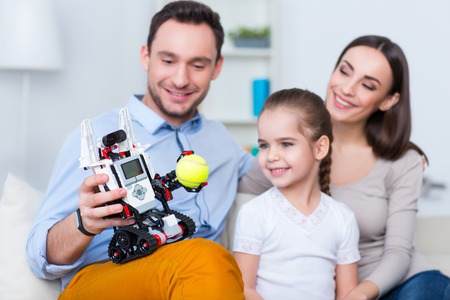 Involved in positivity. Joyful delighted loving family sitting on the couch and playing with robot while resting togetherの写真素材