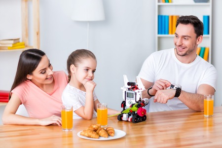 Be in motion. Cheerful delighted smiling family sitting on the floor and eating while playing with robotの写真素材