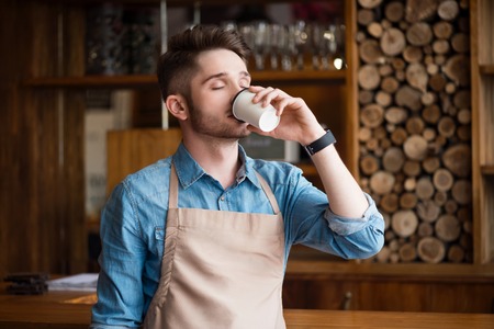 Enjoy the taste. Pleasant content handsome waiter drinking coffee and feeling glad while standing in the cafeの写真素材