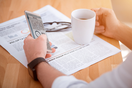 Wave of news. Close up of cell phone  in hands  of pleasant man using it and sitting at the table while drinking coffeeの写真素材