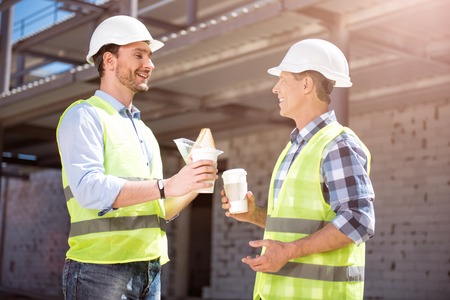 While having a break. Smiling engineers having a break while construction works and eating tasty sandwiches, drinking nice coffee being in a friendly good moodの写真素材