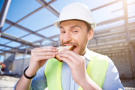 Tasty sandwich.  Smiling and positive builder having lunch and eating tasty sandwiches while being on construction and looking at a cameraの写真素材