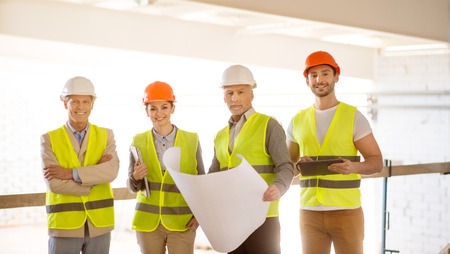 Team spirit. Cheerful and smiling team of architects standing together looking at a camera while holding a new project plan, a digital tablet and some papersの写真素材