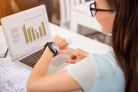 Modern user. Pleasant woman sitting at the table and using her smart watch while working on the laptopの写真素材