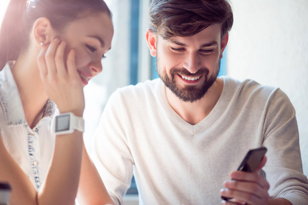 Chatting together. Happy and cheerful modern young woman and man using a smartphone and chatting together sitting in a cafeの写真素材