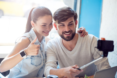 Using a monopod. Happy and merry modern young woman and man sitting in a cafe while drinking coffee and using a smartphone and a selfie stick to take selfie photographの写真素材