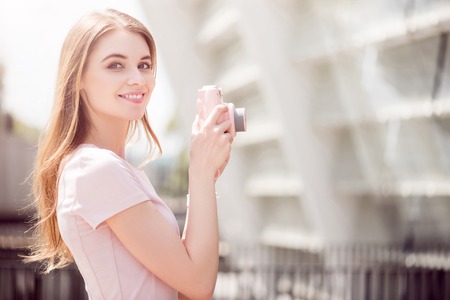 Photo industry. Happy and smiling young woman using her rose camera to take photos while being outdoorsの写真素材