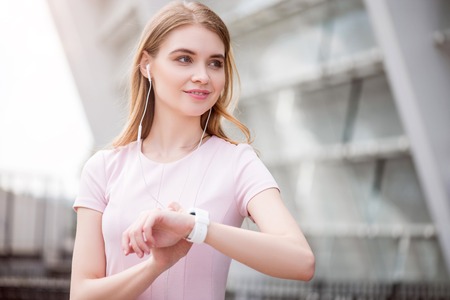 Being modern. Positive and delighted young woman listening to music and checking time on her smart watch while being outdoorsの写真素材