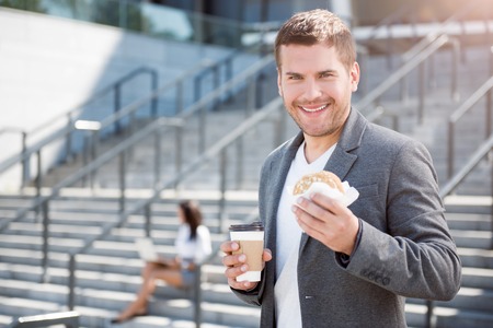 Tasty break. Happy and cheerful young businessman eating sandwich and drinking coffee while a break with his colleague working using a laptop in a backgroundの写真素材