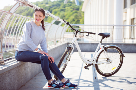 In a good mood. Charming young woman sitting on a border near her bicycle after a good walk and looking at the cameraの写真素材