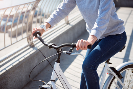 Pleasant day. Close up hands of a young woman wearing a smart watch and holding handles on white bicycleの写真素材
