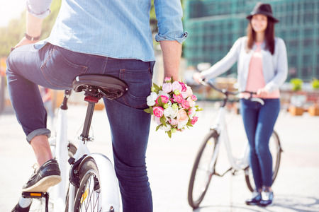 Happy to see her. Picture of legs of a man sitting on a bike and holding flowers with a woman holding a bike on the backgroundの写真素材
